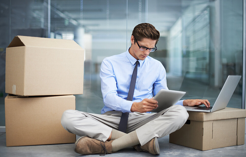 Geschäftsmann sitzt zwischen Umzugskartons, arbeitet mit Tablet und Laptop – Symbolbild für digitale Umzugsorganisation und Bürosoftware.
